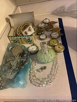 Overview photo showing nine pocket watches arranged on a white surface with costume jewelry including pearl necklaces, aurora borealis crystal necklace, earrings, and decorative green glass perfume bottle.