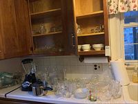 Full view of kitchen counter showing Oster blender, aluminum pot with glass lid, and numerous pieces of glassware including candle holders, small bowls, and glasses.