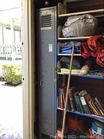Full view of single tall gray metal locker with some surrounding shelving and items on shelves.