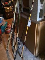 Group of six fly fishing rods standing vertically against a wall, showing cork handles and various colored finishes including natural bamboo and green hues.