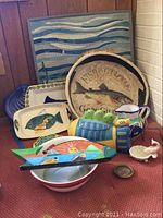 Group of multiple fish themed ceramic platters, bowls, cookie jar, pitcher, and wall art arranged on red carpeted surface against wood panel wall