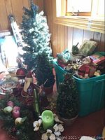Overall lot showing two faux tabletop Christmas trees in planters with surrounding holiday decor and a green plastic bin containing additional holiday items.