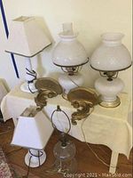 Photo showing entire lot with lamps and sconces arranged on a table. Two white fabric shade lamps, two milk glass lamps with hobnail pattern, one missing chimney shade, glass base lamp without shade, and two gold-toned wall sconces visible.