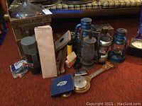 Wide view showing glass carboy in wooden frame, several lanterns, portable drink dispenser in packaging, barometer, binoculars, flashlight, suspenders, and two metal lighters.