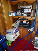 Photo showing wooden shelf corner with stacked boxes including Custom Electric Slicing Knife, Chef's Choice sharpener box, playing cards, and various kitchen tools and knives visible. Items dusty.