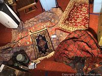 Photo showing multiple rugs piled on an orange carpet, including a large rust-toned decorative rug and smaller rugs.