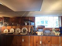 Wide view showing an assortment of items including several decorative plates, cut glass serving pieces and bowls, crystal vases and glasses arranged on shelving under window with wood paneling background.