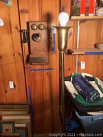 Photo showing entire antique wooden wall telephone mounted on wooden paneled wall next to brass floor lamp without shade