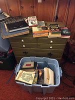 Assortment of books on green dresser and in blue plastic bin