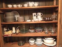 Photo showing shelves with various dishes including white soup crocks, plates, coffee cups, glass bowls, and glass tumblers.