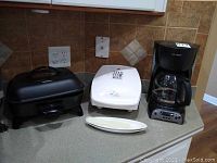 Photo showing George Foreman grill, Mr. Coffee maker, and Rival deep fryer side by side on kitchen countertop, visible wear and scratches on appliances.