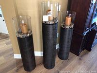 Overall view of three tower-style candle holders made of dark textured metal bases and clear acrylic tops containing brown pillar candles and stones, showing size variations and design.