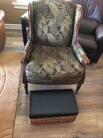 Front view of the carved wood arm chair with leaf-pattern upholstery and vinyl-covered raffia foot stool.