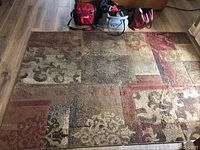Top view of rectangular floor rug showing multicolored patchwork design with floral and leaf motifs in brown, red, beige tones on wood flooring.