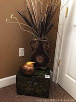 Photo showing metal trunk at the bottom, wooden plant holder with faux plants above it, and ceramic bird on top of trunk in corner setting.