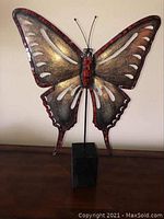 Front view of metal butterfly wall art with wood stand, showing painted details and glass mosaic edges with some missing pieces.