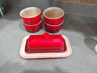Photo showing one Le Creuset butter dish with lid and four red and cream ramekins on kitchen counter.