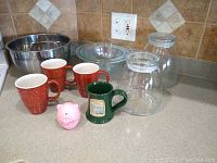 Set of three graduated Pyrex bowls, three textured orange Pier One mugs, one green Harrington House ceramic mug, two clear glass canisters, pink pig timer, and a stainless steel mixing bowl all displayed on kitchen counter.
