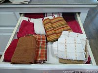 Drawer with assorted folded kitchen dish towels and dish rags showing various colors and patterns including red, white, orange, and brown