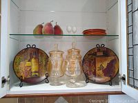 Shelved display showing decorative plates with grape and wine motifs, three clear glass apothecary jars, two small white egg-shaped objects, three artificial pears, and stacked orange Spectrum plates.