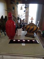 Photo showing large red urn, ornate pedestal urn, white vase with peacock feathers, and red candle tray on countertop