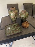 Three ceramic decor pieces on a glass table: two vases and one platter, all decorated with green floral motifs on a brown background. Also visible is a large piece of rope beside the platter.