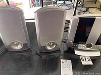 Photo of grey CD player with alarm clock surrounded by two standing speakers on each side and remote on the table in front.