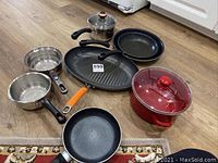 Overview of all pots and pans including red covered pot, frying pans, and two aluminum saucepans on wooden floor