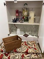 Open cabinet showcasing an assortment of bar-related glassware, ceramic ice canister, basket, and patterned fabric items spread out on the cabinet shelf and bottom.