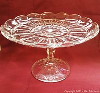 Clear glass cake plate on a stemmed pedestal with a scalloped edge and decorative pattern.
