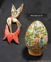 Photo showing metal decorative egg with colorful bird motifs next to wood carved painted hummingbird