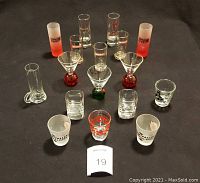 Photo showing 17 shot glasses arranged on a black surface, various shapes including tall, cylindrical, frosted, colored stem and square base styles.