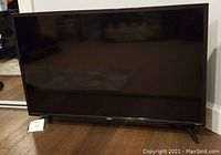 Front view of the 36-inch RCA Roku TV showing the TV screen, black glossy bezel, and RCA logo at center bottom. The TV is resting on a hardwood floor with reflections visible on the screen.