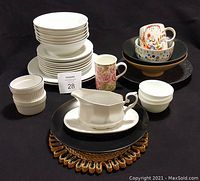 Photo showing multiple stacks of various dishes including white plates with a blue rim, black plates, floral patterned cups and mugs, white gravy boat, ramekins, and covered bowls.