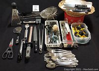 Wide view of assortment of cutlery and kitchen utensils including steak knives, garlic presses, grater, cake decorating kit, and serving spoons all arranged on a black surface.