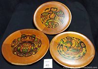Three vintage round wooden plates with colorful Northwest Native American style artwork on front, set on black background.