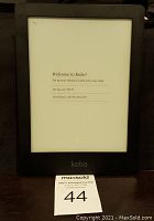 Frontal photo of the Kobo eReader powered on, showing a welcome setup screen. Device is black with visible 'kobo' logo at bottom bezel.
