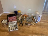 Wide photo showing all baking and kitchen items arranged on hardwood floor: muffin tins, trays, pitchers, can opener, silverware sets and holders, dish drying rack, rolling pin, pizza cutter, measuring cup, and assorted utensils.