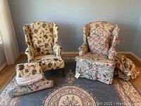 Pair of matching wing back chairs upholstered in cream embroidered floral fabric with deep pink velvet backs. Both chairs include matching cushions. Chairs resting on floral patterned rug in front of pale blue wall.