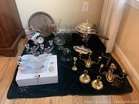 Photo displaying assortment of silver plated, brass, porcelain, and glass items arranged on black cloth including chafing dish, butter dish, candlesticks, porcelain dishes, decorative glass pieces, and clock.