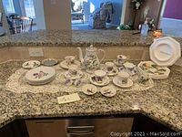 Overall collection of fine china tea cups, saucers, plates, and a teapot displayed on a kitchen counter with a lace table runner