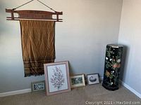 Full lot view showing the textile wall hanging on the wall, four framed pictures on the floor, and the black octagonal decorative furniture piece in the corner