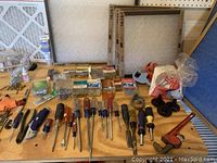 Photo showing an assortment of used hand tools including screwdrivers, pliers, a pipe wrench, screws, fasteners, and new air filters on a wooden table.