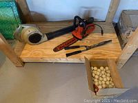 Photo showing the lot contents including an orange and black electric chainsaw, beige leaf blower, manual hedge trimmers, and a box of wooden balls on a wooden shelf.