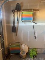 Wide shot of all items hanging on wall or resting on floor including two shovels, beach chairs, ceramic pot, weighted umbrella base, and lawn hook.