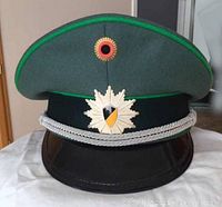 Front view of the green police hat showing the white starburst badge with black and gold Baden-Württemberg shield, silver chin cord, green piping, black glossy visor, and pebble side buttons.