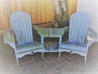 Front view showing two blue-painted wooden Muskoka chairs attached by a small triangular table, outdoors on a stone patio with a wooden fence and tree in the background. Mossy algae visible on wood surfaces.