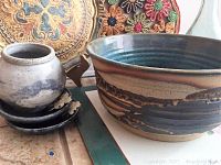 Close-up of large ceramic bowl with blue-green interior and brown exterior glaze, alongside small ceramic pot and decorative coaster, metal plate visible in background.