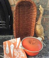 Photo showing the orange Le Creuset pot with lid, wicker basket behind it, orange printed tablecloth, and duck decoration to the right.