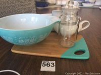 Photo showing large aqua blue Pyrex Amish Butterprint Cinderella bowl, white cream jars, glass jar with lid, and bamboo cutting board with turquoise corner detail.